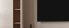 Warm minimalist living room feature wall with wall-mounted TV, vertical open display shelves, low media console, and decorative ceramic vase.