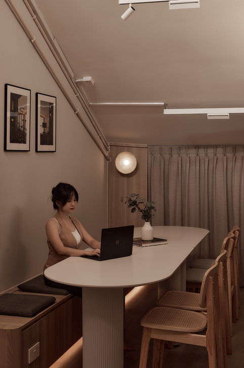 A woman working on a laptop at a long, curved white communal table in a warm, minimalist studio setting with light oak accents.