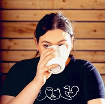 A white person with tied-back brown hair raises a cup of tea or coffee to their mouth and drinks. They are wearing a dark blue t-shirt with a white illustration on the front and there is wooden slats on the wall in the background.