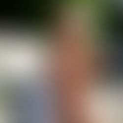 Woman in a white dress sits on a bench, smiling and holding an iced drink. Sunlight filters through trees, creating a relaxed mood.