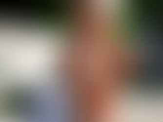 Woman in a white dress sits on a bench, smiling and holding an iced drink. Sunlight filters through trees, creating a relaxed mood.