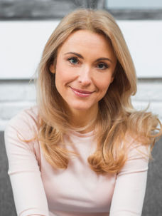 Smiling woman with blonde hair wearing a light pink top, seated on a gray sofa. White brick wall background, bright and inviting mood.