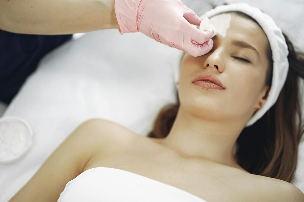 A woman with eyes closed receives a facial treatment. Pink-gloved hand applies cream on her forehead. She wears a white headband. Relaxed mood.