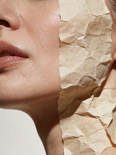Close-up of a face partially covered by crumpled beige paper, highlighting smooth skin and glossy lips. Soft lighting creates a serene mood.