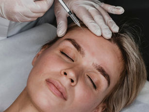 Close-up of a person receiving a cosmetic injection in the forehead. Eyes closed, relaxed on a white pillow. Gloved hands are visible.