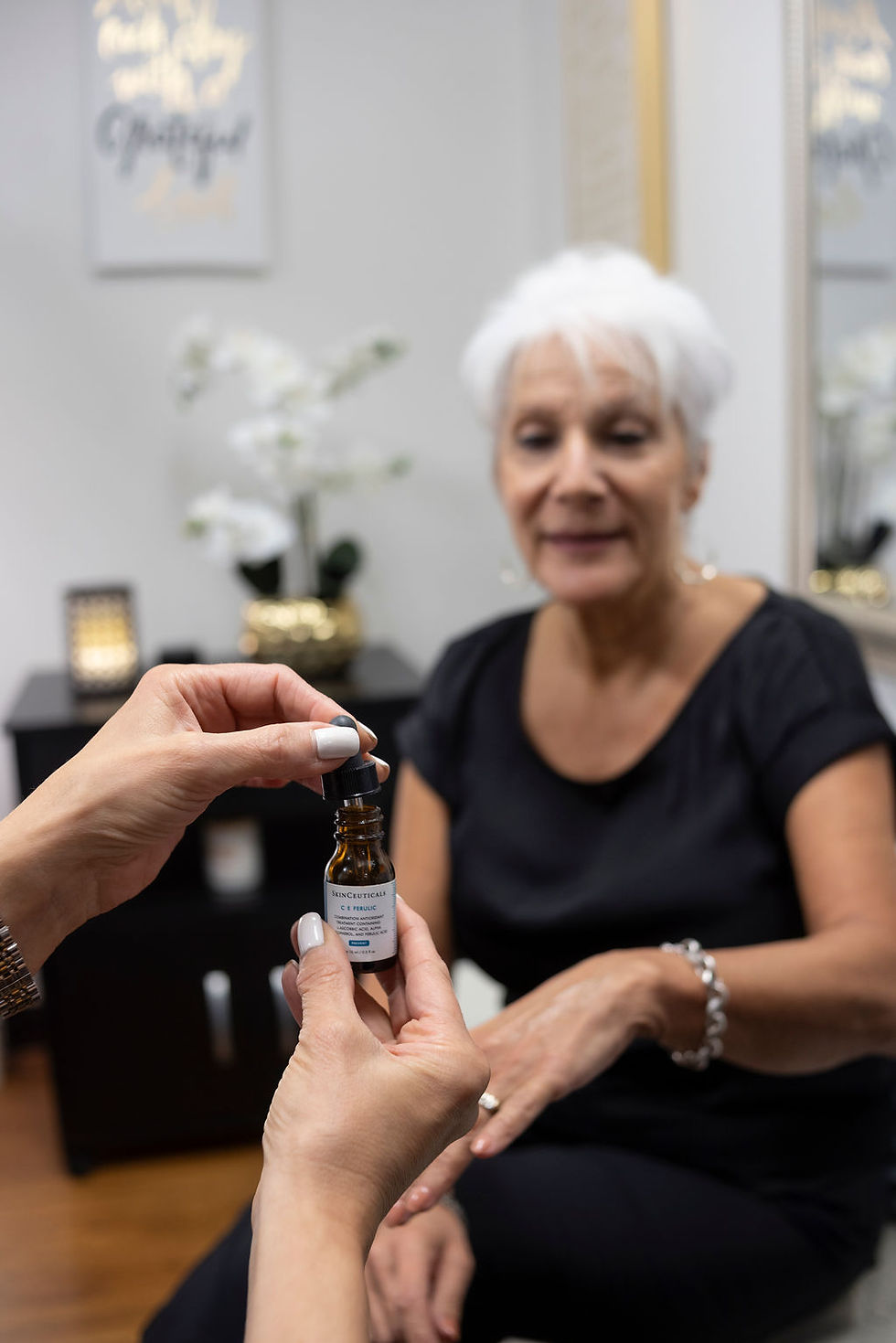 Hands hold a SkinCeuticals C E Ferulic serum bottle before an older woman in a calm spa room.