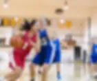 A girl in a red jersey dribbles a basketball, pursued by opponents in blue jerseys on an indoor court. Background banners and bright lights.
