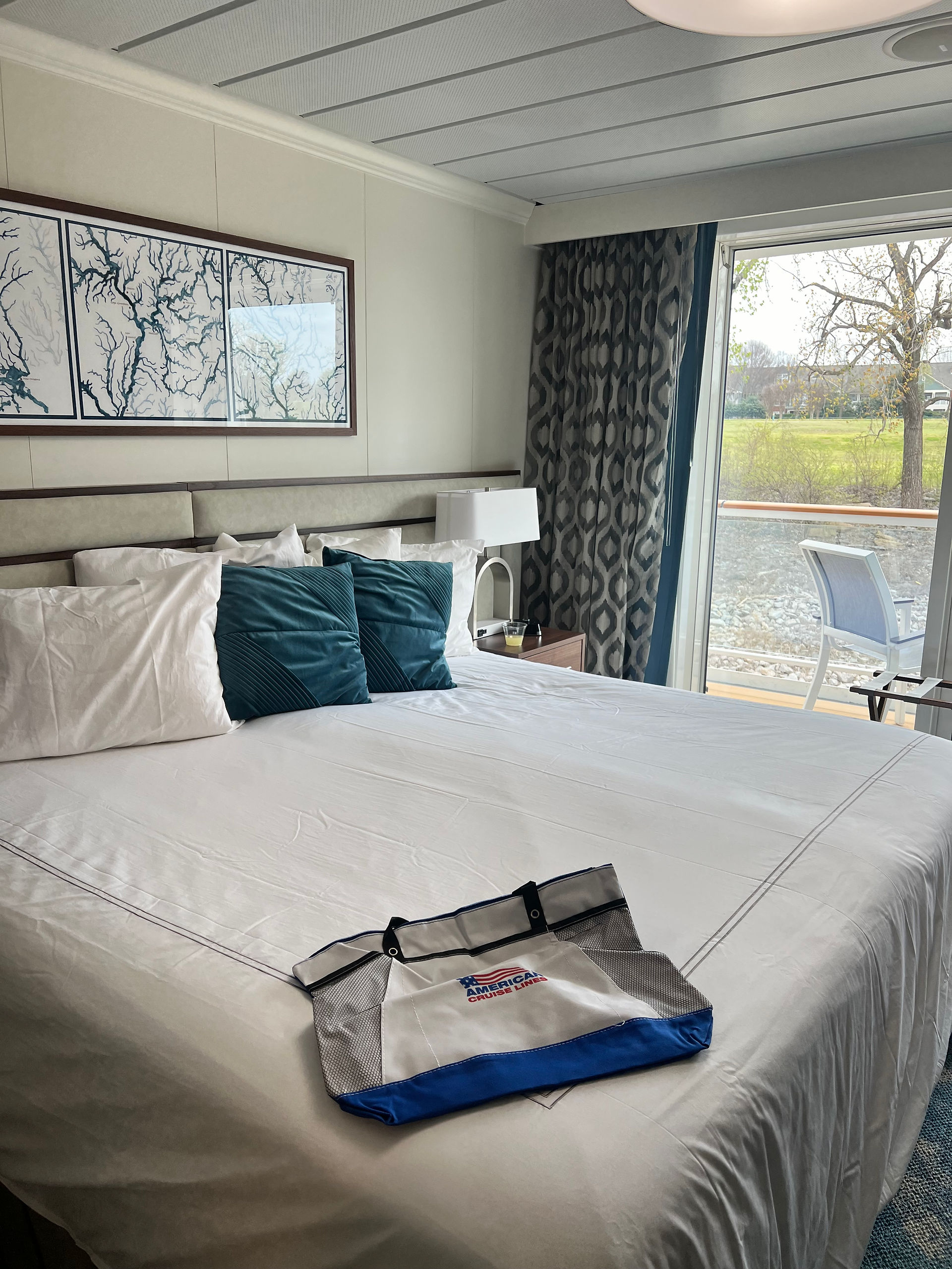 Photo of the bed and balcony of a balcony stateroom.   The room is mostly blue and white and the bed is a king side bed.  The balcony is big enough for 2 chairs and a table.