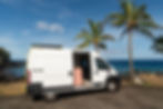 Laverne, a pink glampervan with Sun + Salt Campervans parked next to some palm trees on pavement by the side of the beautiful blue ocean in Hawaii.