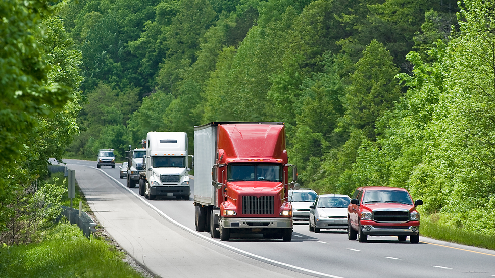 Red truck leads cars and other trucks on a highway lined with lush green trees, under a clear sky, conveying a busy, vibrant scene.