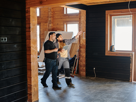 Two people discussing home renovation inside a wooden house. They examine a black wood-paneled wall, holding a tablet. Bright window light.
