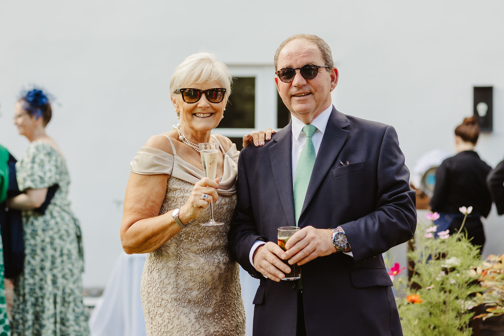 2 wedding guests, husband and wife smile at the camera in wedding attire and sungalsses