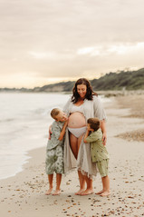 2 brothers cuddle their pregnant mum on the beach. Mum wears a white lace kaftan showing her bump