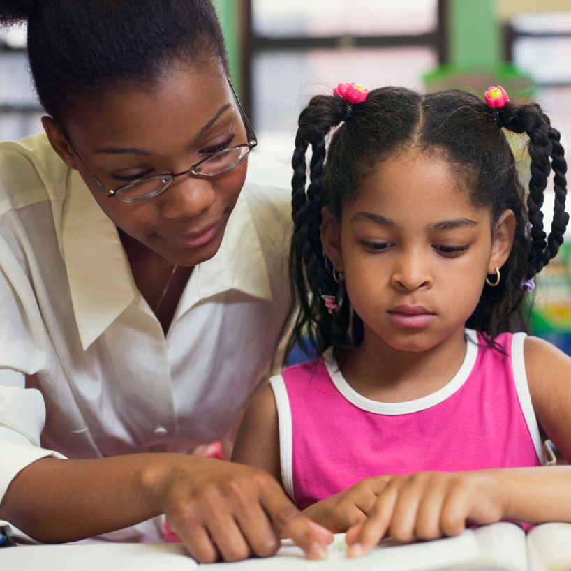 Therapist helps a focused young girl read a book in a classroom. The teacher points to the text. The girl wears a pink dress, and her hair is braided.