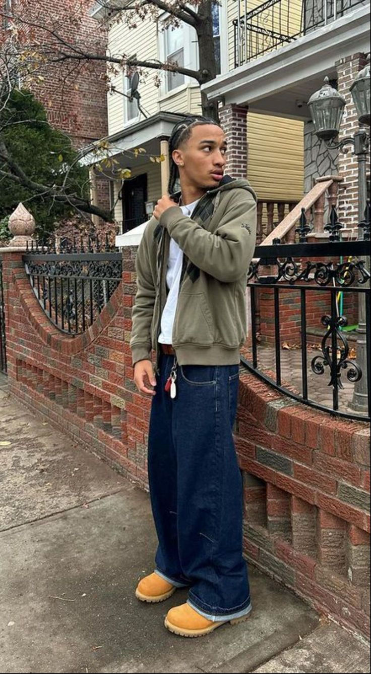 Man wearing a casual streetwear outfit with an olive jacket, white shirt, baggy jeans, and Timberland boots, standing on a city sidewalk in front of a brick building.