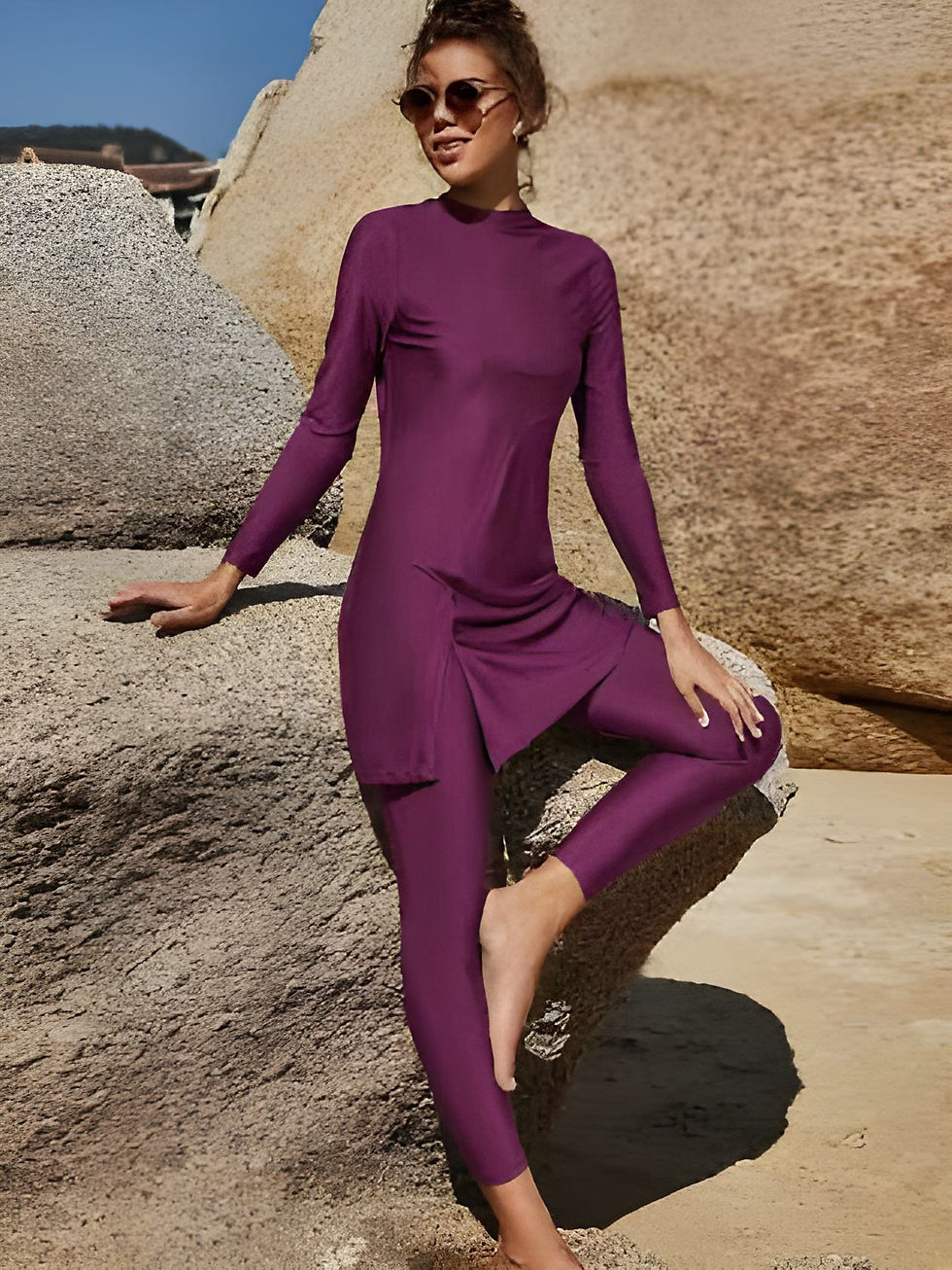 Woman wearing a long-sleeved, full-length purple burkini with leggings, sitting on rocks at the beach and wearing sunglasses.