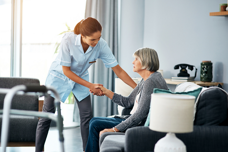 A COMPASSIONATE HOME HEALTHCARE AIDE SHAKING HANDS AND HELPING AN ELDERLY WOMAN ON HER LIVING ROOM COUCH.