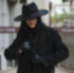 a woman in a black trench coat with a wide brim black hat and leather gloves.