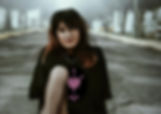 A woman in fishnets sitting on the ground in a cemetery wearing a black t-shirt with a pink heart pierced with three swords on it.