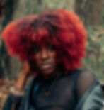 A black woman with red hair color and dark roots wearing a black mesh top and a leather jacket.