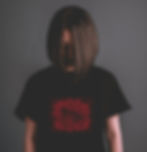 A woman in a black t-shirt with a red wolf on it standing in front of a dark background with her head hanging down.