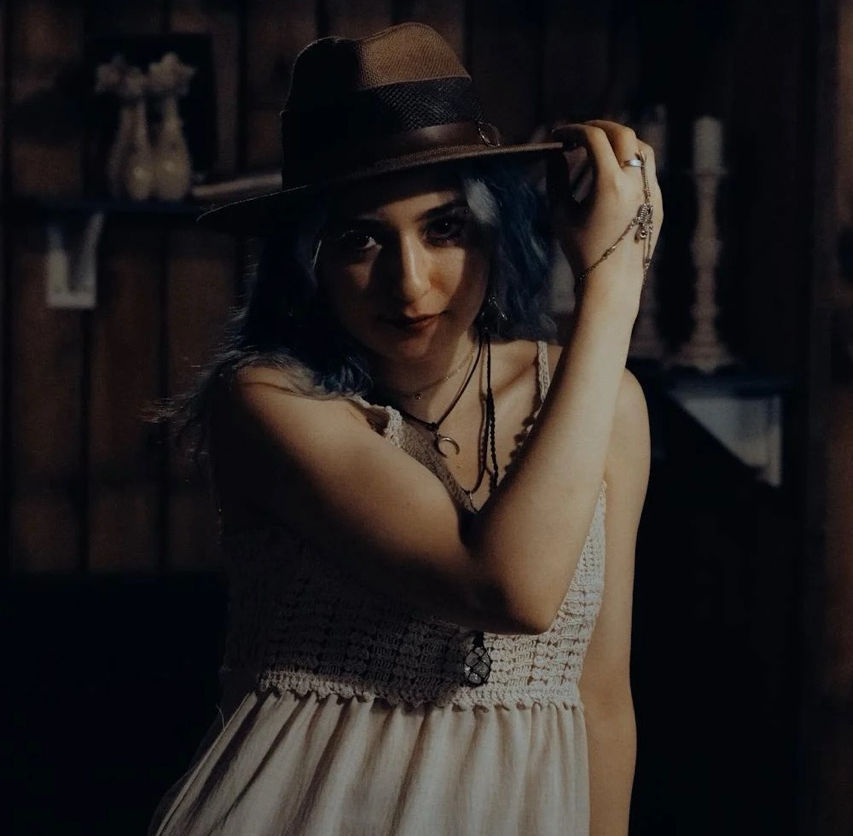 a woman in a beige sundress with a brown hat wearing several black necklaces and bracelets holding her hat brim