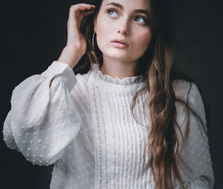 a woman wearing a lacy white long sleeve blouse looking away to the left on a black background.