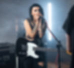 A woman with black hair and tattoos holding a guitar singing into a microphone.