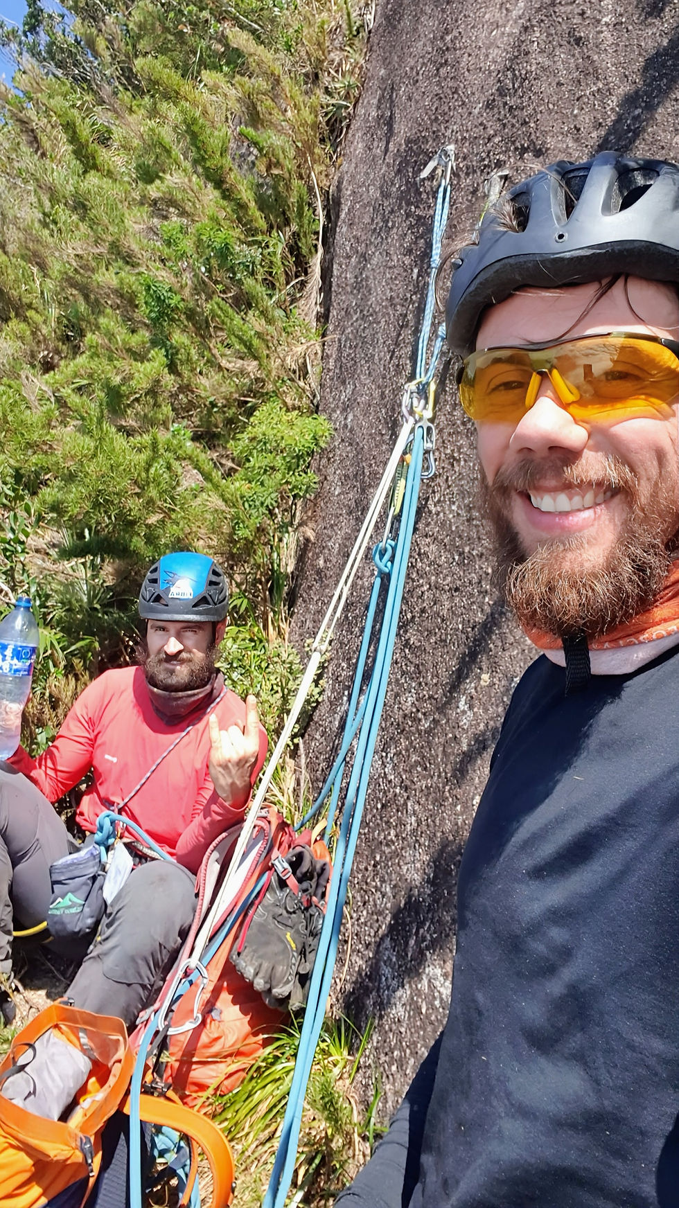 Dois escaladores com capacetes e equipamentos de segurança estão ancorados a uma parede de rocha, sorrindo e descansando em meio à vegetação de montanha. Um deles segura uma garrafa de água.
