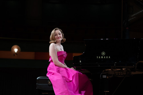 Pianist in a pink dress smiles near a Yamaha piano in a hall.