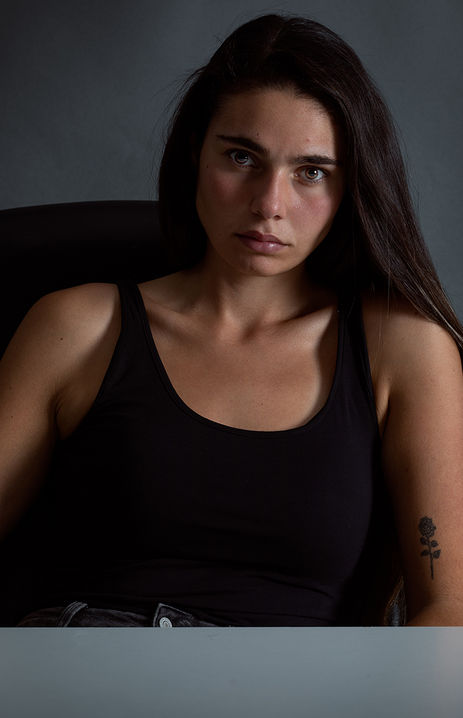 "Female model Angie Alvarez sitting on a black chair wearing a basic black tank top against a dark gray background in a studio setting."