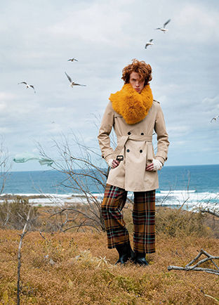 Full-length color portrait of redhair male model Nelson Del Toro posing elegant in a cold coastal field. Wearing a double-breasted coat, yellow fur scarf, and plaid trousers, with seagulls flying in a cloudy sky.