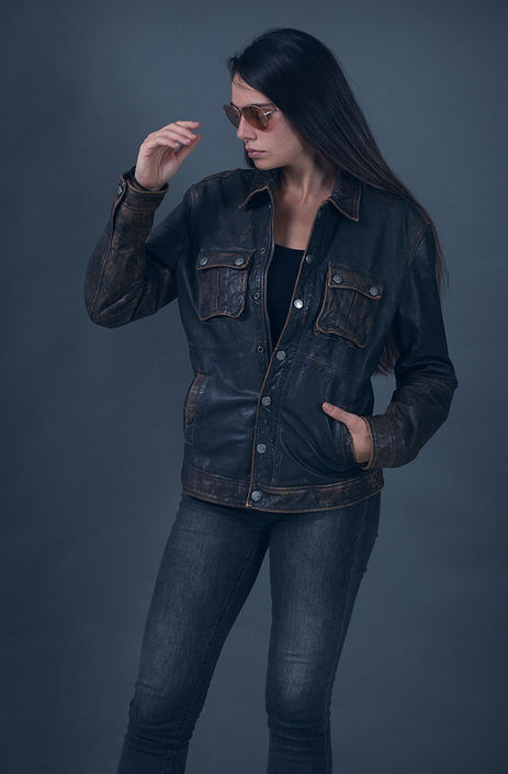"Fashion model Angie Alvarez wearing a distressed brown leather Armani jacket and pink aviator sunglasses in a studio setting against a gray background."
