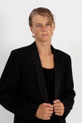 Studio photograph of male model Neo Rosdahl wearing a black tuxedo jacket over a black tank top and fitted black jeans against a clean white background.