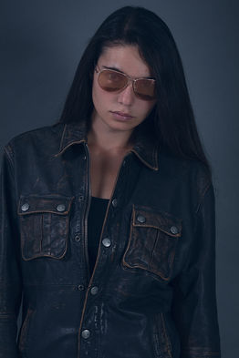 "Fashion model Angie Alvarez wearing a distressed brown leather Armani jacket and pink aviator sunglasses in a studio setting against a gray background."
