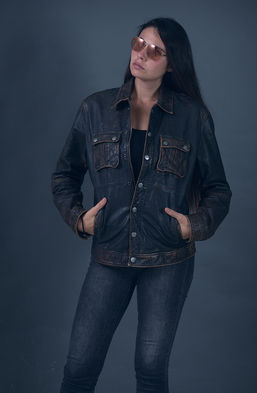 "Fashion model Angie Alvarez wearing a distressed brown leather Armani jacket and pink aviator sunglasses in a studio setting against a gray background."