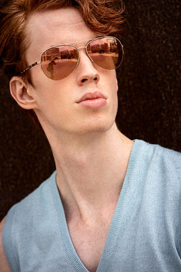 Outdoor photograph of redhair male model Nelson Del Toro wearing a cream sleeveless v-neck sweater with no shirt and aviator sunglasses that match his hair color.