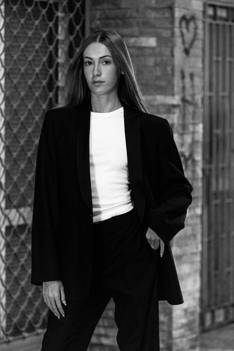 Outdoor portrait of female model Sara Jimenez wearing a black tuxedo jacket and trousers over a white tank top against a neutral background.