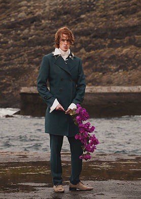 Full-length photograph of redhair male model Nelson Del Toro posing on a coast. Wearing a bottle green suit and overcoat, an oversized white collar shirt with folded big cuffs, holding long purple flowers in his left hand.