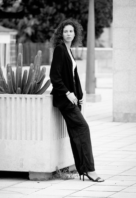 Black and white street fashion portrait of model Mireya Garcia posing relaxed against a wall with Las Palmas de Gran Canaria city background, wearing a white tank top and black jacket.