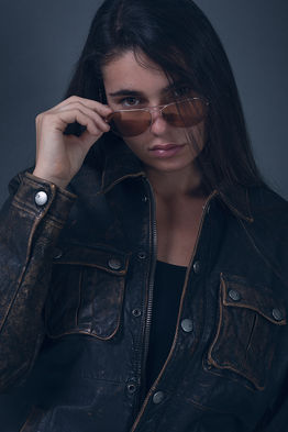 "Fashion model Angie Alvarez wearing a distressed brown leather Armani jacket and pink aviator sunglasses in a studio setting against a gray background."