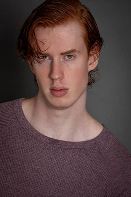 Studio portrait of redhair male model Nelson Del Toro against a grey background. Wearing a burgundy crewneck sweater with his hair slicked back and styled to the side.