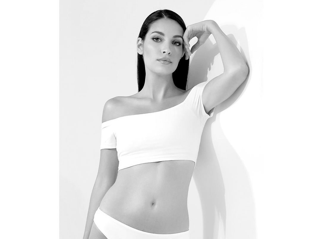 Runway model Lourdes López (182 cm) posing for official agency digitals. Clean studio photoshoot with a bright background, wearing a basic white camisole and white briefs. Natural profile for international high-fashion campaigns.