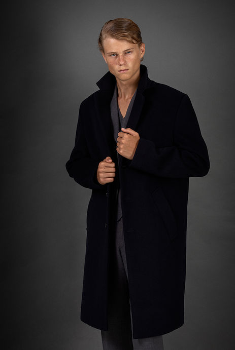 Studio fashion photograph of male model Neo Rosdahl wearing an elegant black three-quarter overcoat over a tailored suit against a grey background.