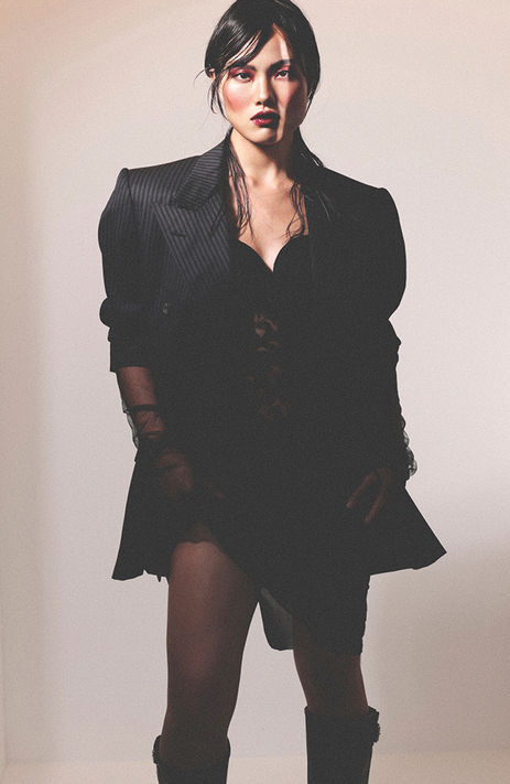 Studio fashion photograph of East Asian female model Giulia Xie wearing a black nightgown, a dark jacket, sheer dark tights, and high boots, featuring bold dramatic makeup and a sleek hair updo.
