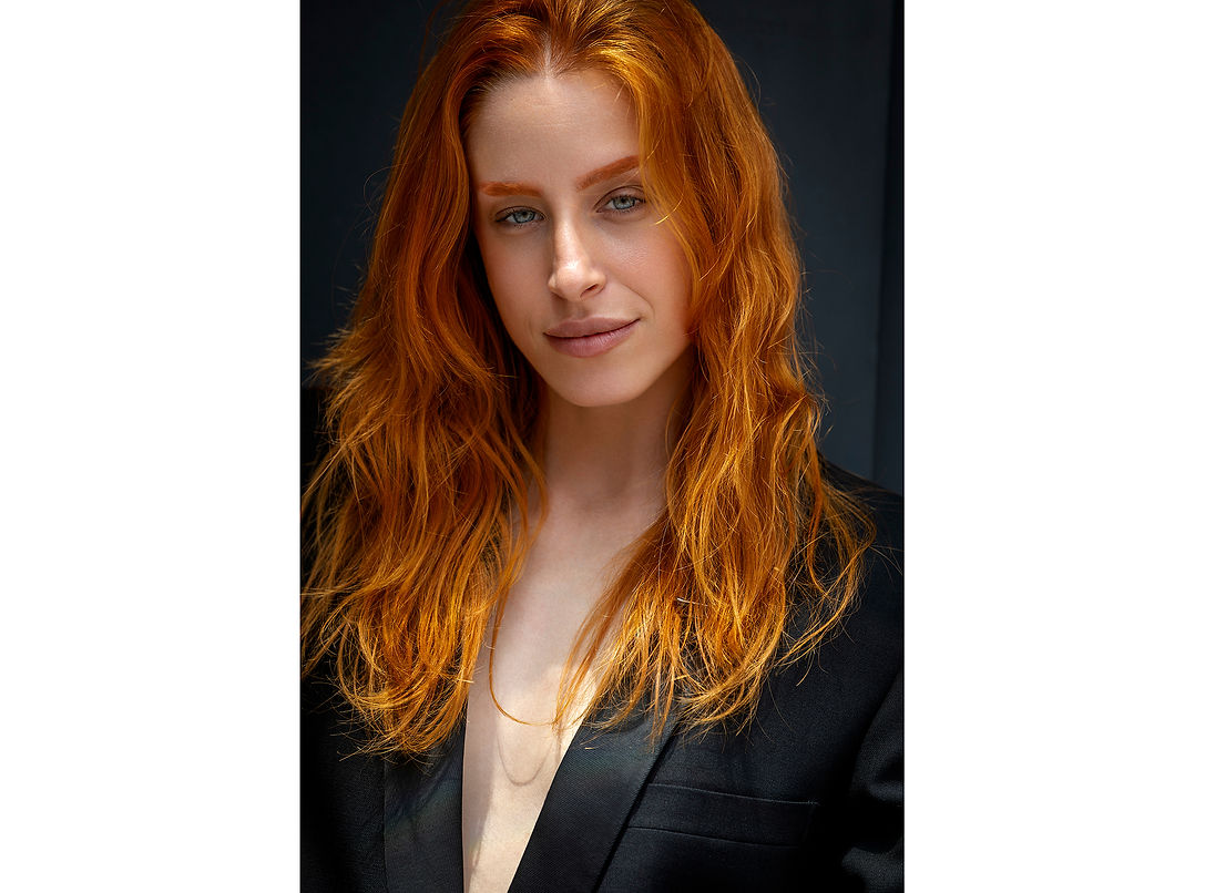 A striking and sophisticated close-up portrait of redhead female model Elena Vázquez. She is wearing an elegant black blazer against a solid black background, creating a high-contrast look that beautifully emphasizes her bright red hair, natural skin tone, and facial features under professional studio lighting.