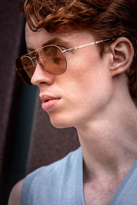 Outdoor photograph of redhair male model Nelson Del Toro wearing a cream sleeveless v-neck sweater with no shirt and aviator sunglasses that match his hair color.