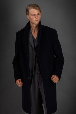 Studio fashion photograph of male model Neo Rosdahl wearing an elegant black three-quarter overcoat over a tailored suit against a grey background.