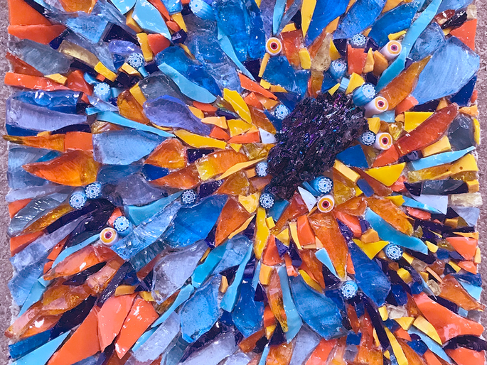 Mosaïque carrée aux couleurs vives organisées en composition radiale évoquant une fleur. Un centre sombre et texturé est entouré d'éclats de verre bleus (roi, ciel, turquoise), orange vif, jaune et noir, qui rayonnent vers les bords. De petites perles décoratives avec motifs circulaires sont dispersées dans la composition. Les fragments de verre de tailles et formes variées créent un relief important et une surface brillante et réfléchissante. L'assemblage dynamique suit un mouvement circulaire depuis le centre. Aucune bordure ou cadre visible sur l'image.