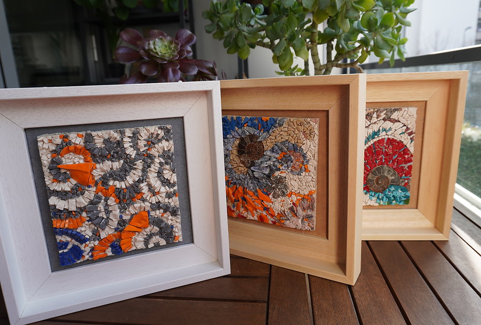 Photographie présentant trois tableaux de mosaïque carrés alignés et encadrés, vus en perspective. Chaque œuvre assemble des tesselles de couleurs vives et de matières variées créant un relief texturé. À gauche, motifs abstraits orange, gris anthracite et blanc sur fond sombre. Au centre et à droite, compositions intégrant des fossiles d'ammonites spiralés entourés de fragments orange, bleu, rouge et turquoise. Les cadres varient entre bois clair et blanc. Technique de mosaïque visible par l'assemblage de petits fragments irréguliers.